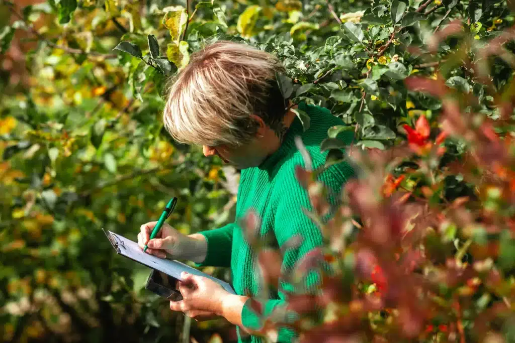 Cornelia schreibt umrahmt von Blättern und Sträuchern auf ihr Klemmbrett.
