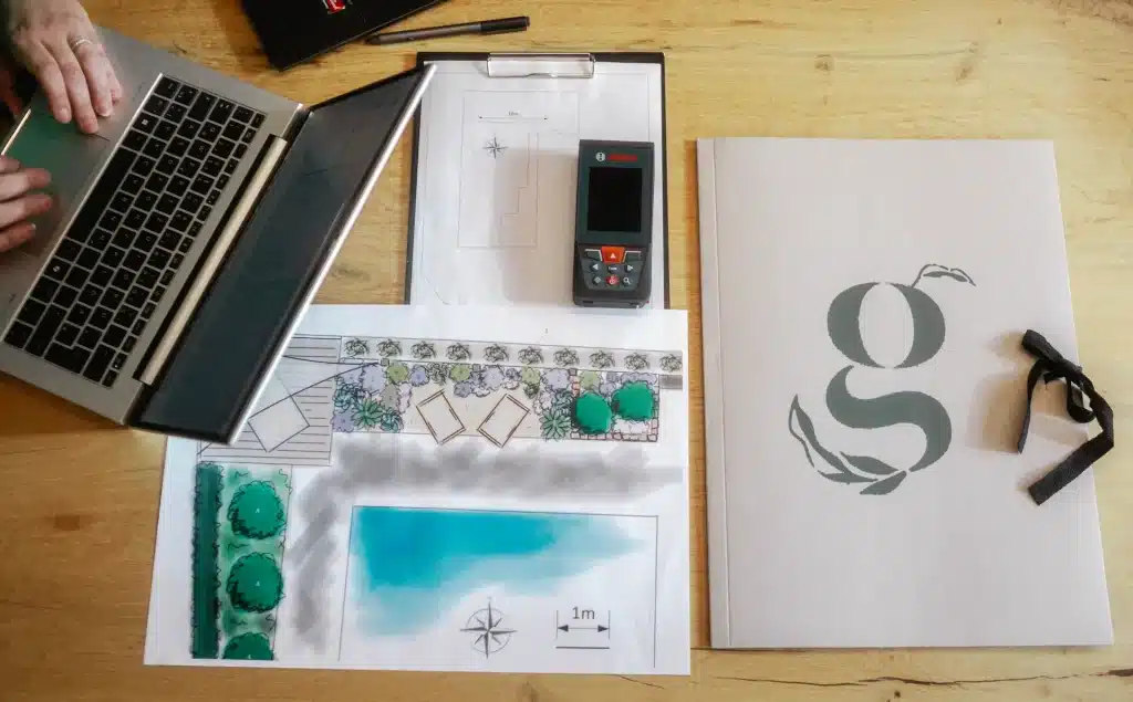 Blick von oben auf einen Schreibtisch auf dem ein Plan, eine Kundenmappe, ein Skizzenblock und ein Laptop stehen.