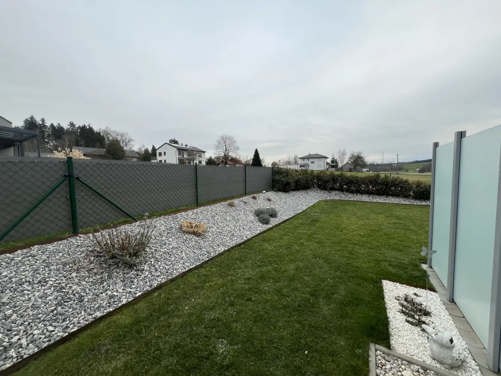 Vorher-Ansicht des Gartenprojekts Klingersberger. Dunkler Zaun umrahmt ein Kiesbeet. In der Mitte eine längliche Rasenfläche.